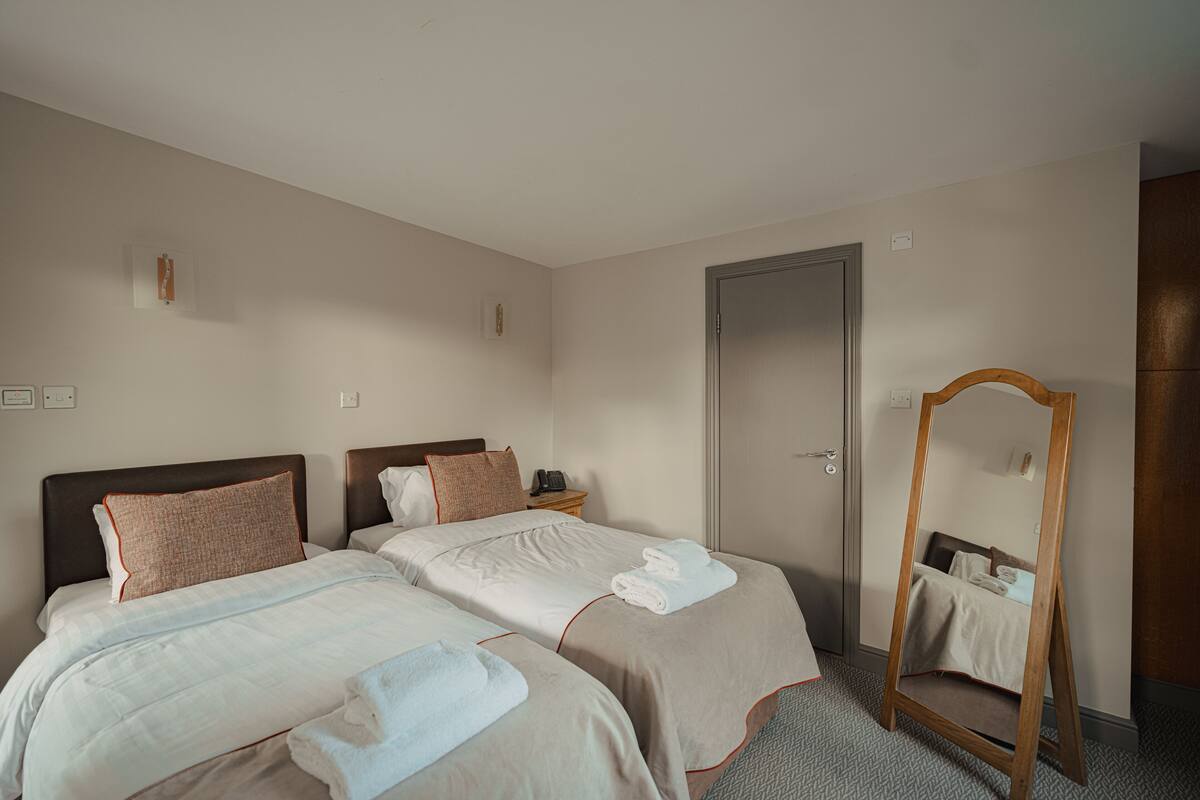 A hotel room with two beds, a wooden-framed mirror, a door, and a small table with a telephone.