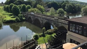 Un puente de piedra sobre un río, con abundante vegetación y un edificio con tejado inclinado al frente.