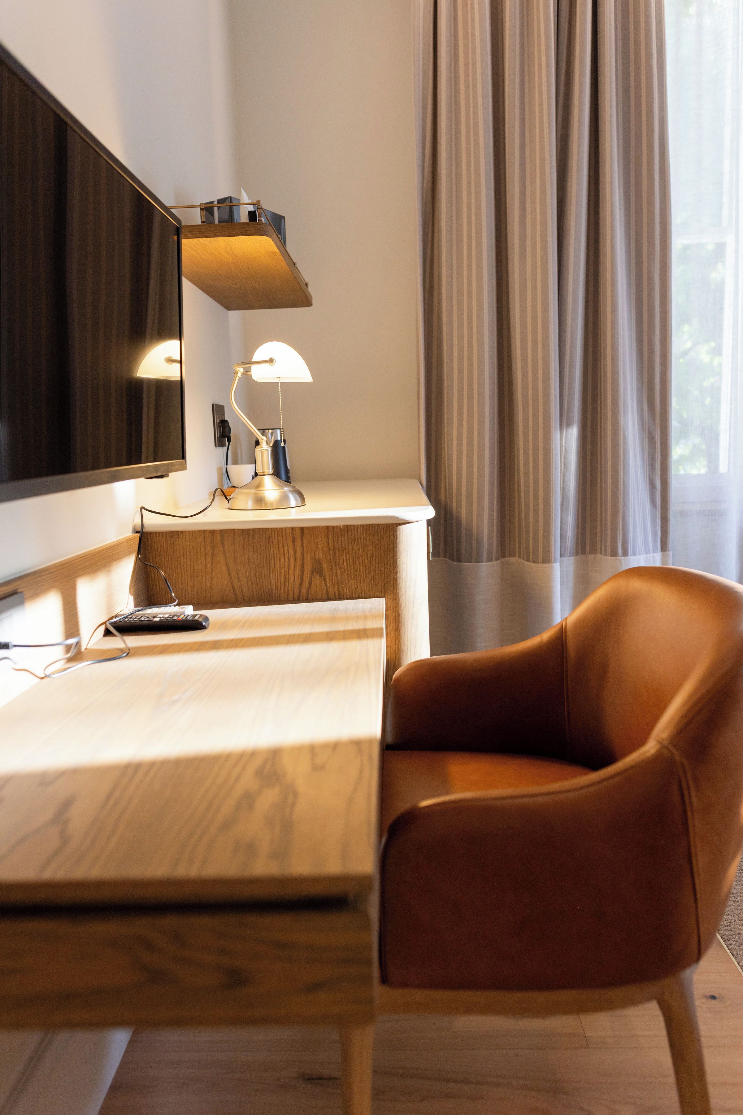 Une chambre d’hôtel moderne, dotée d’un bureau en bois, d’un fauteuil en cuir, d’un téléviseur à écran plat fixé au mur et d’une fenêtre avec des rideaux.
