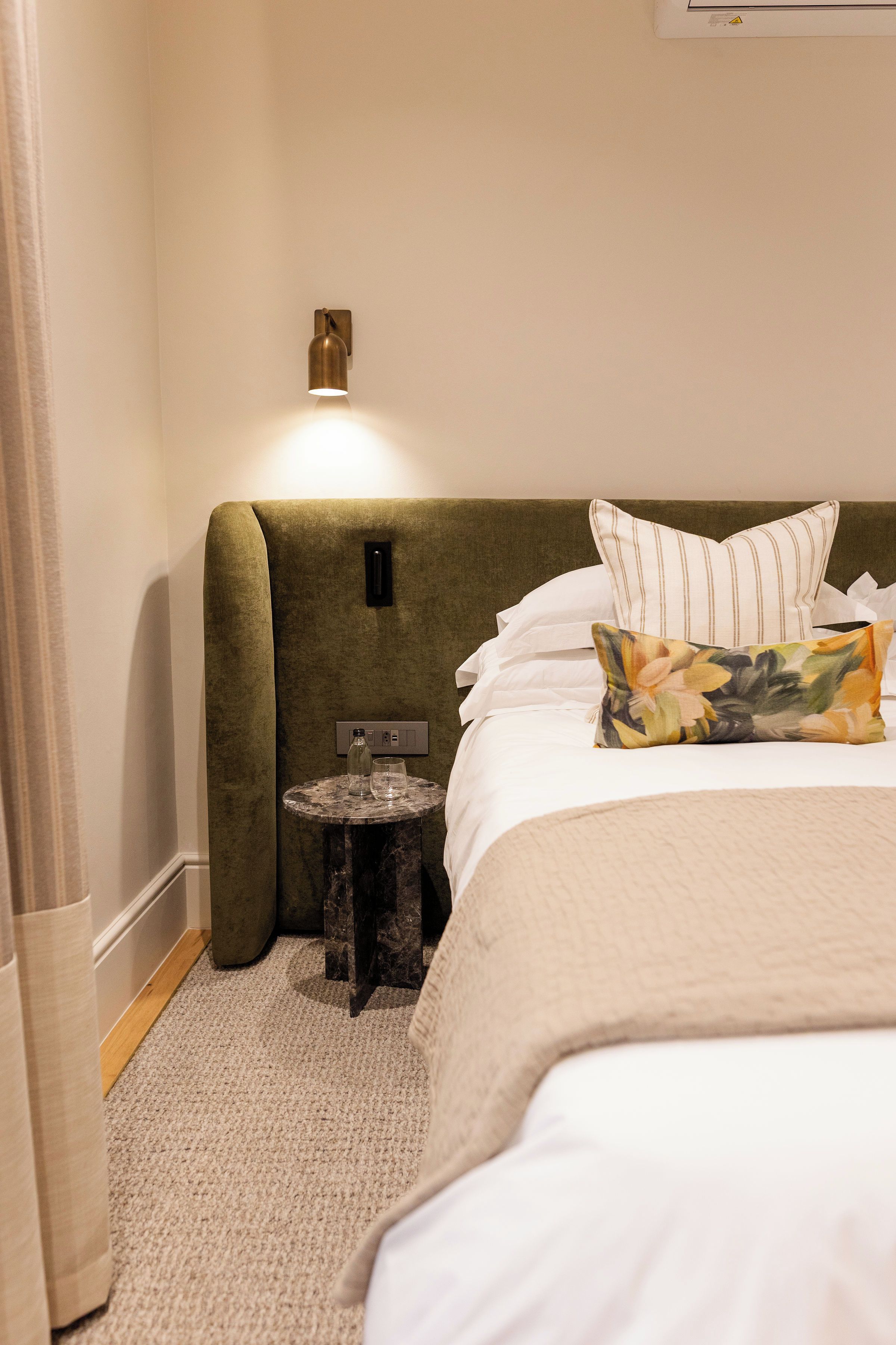 Quarto de hotel com cama, mesa de cabeceira, abajur e uma pequena mesa redonda com garrafa e copo.