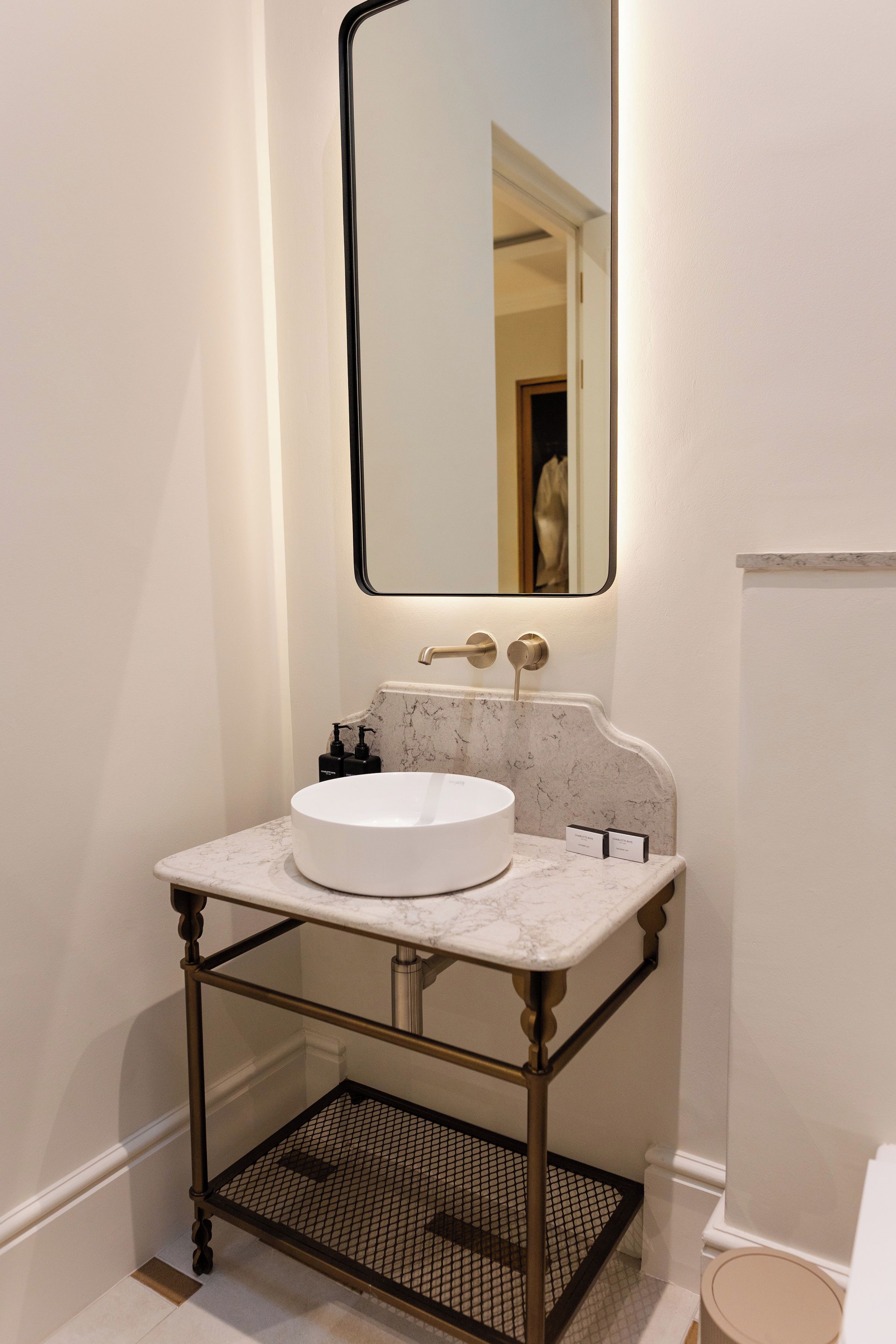 Un baño con lavamanos blanco, encimera de mármol, espejo con marco negro y un estante con artículos de tocador.