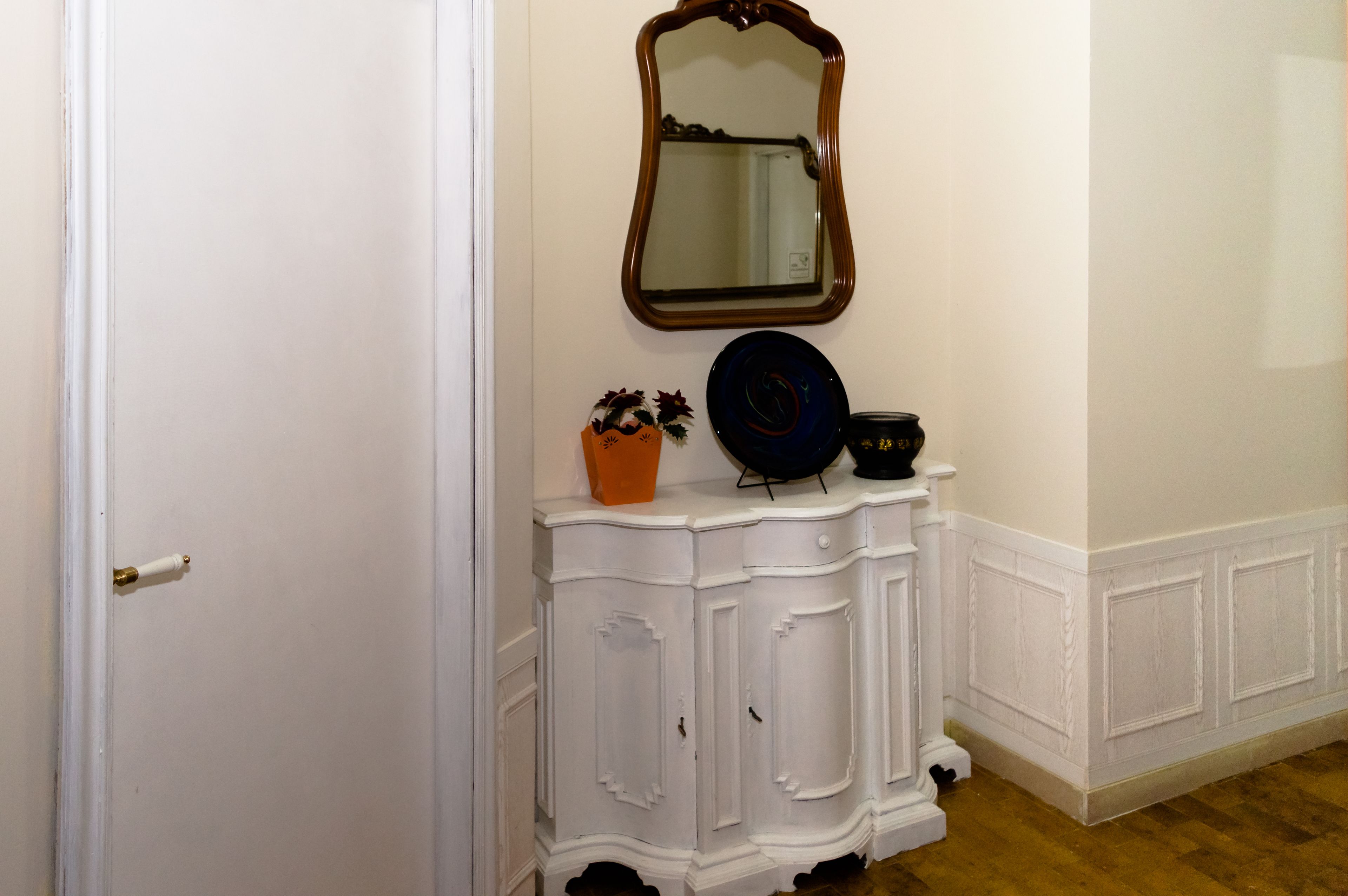 Un mueble blanco con un espejo encima, un pequeño florero naranja con una planta, y un cuenco negro sobre el mueble.