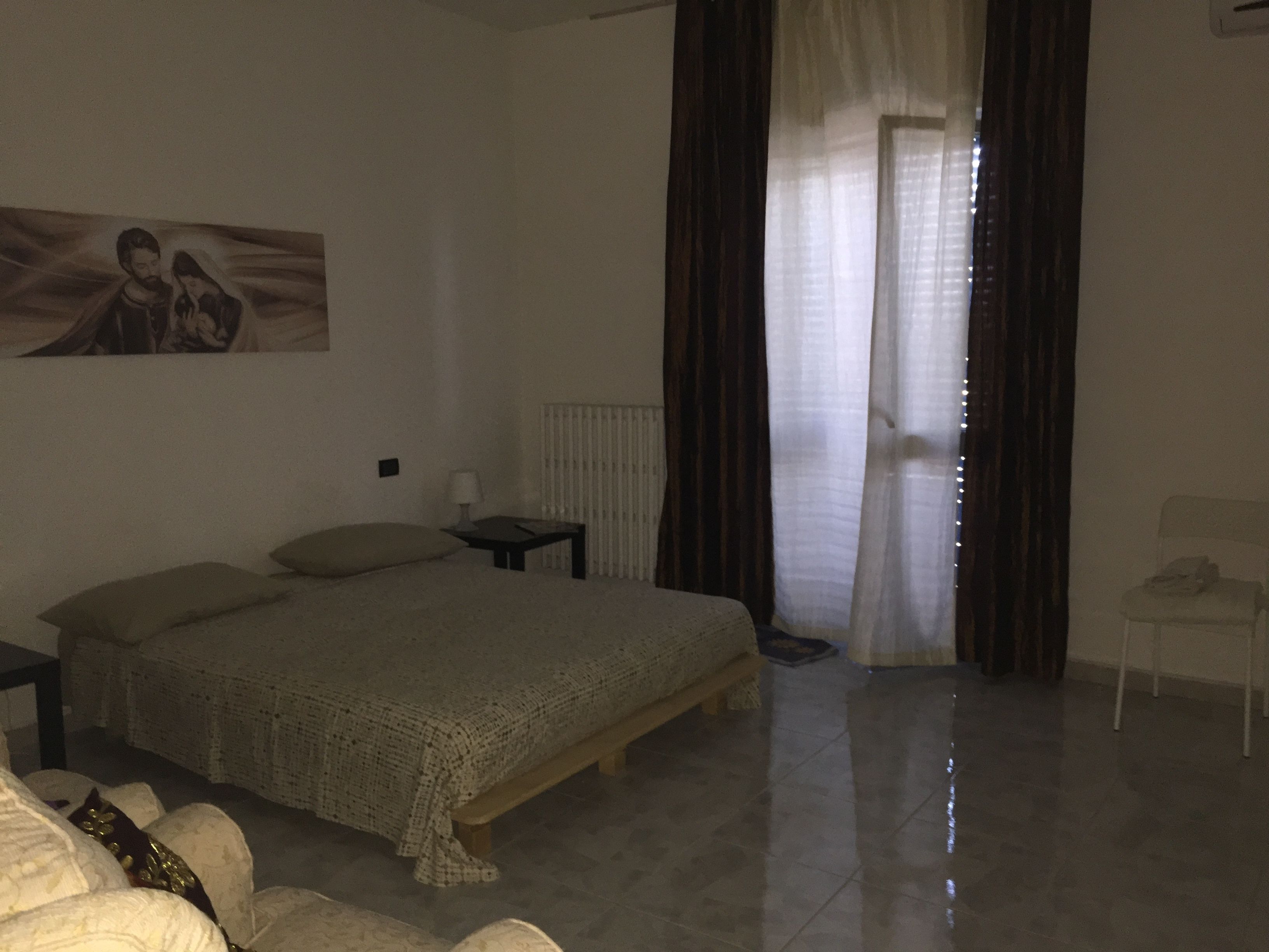 A bedroom with a bed, a nightstand, a chair, a radiator, and a window with curtains.