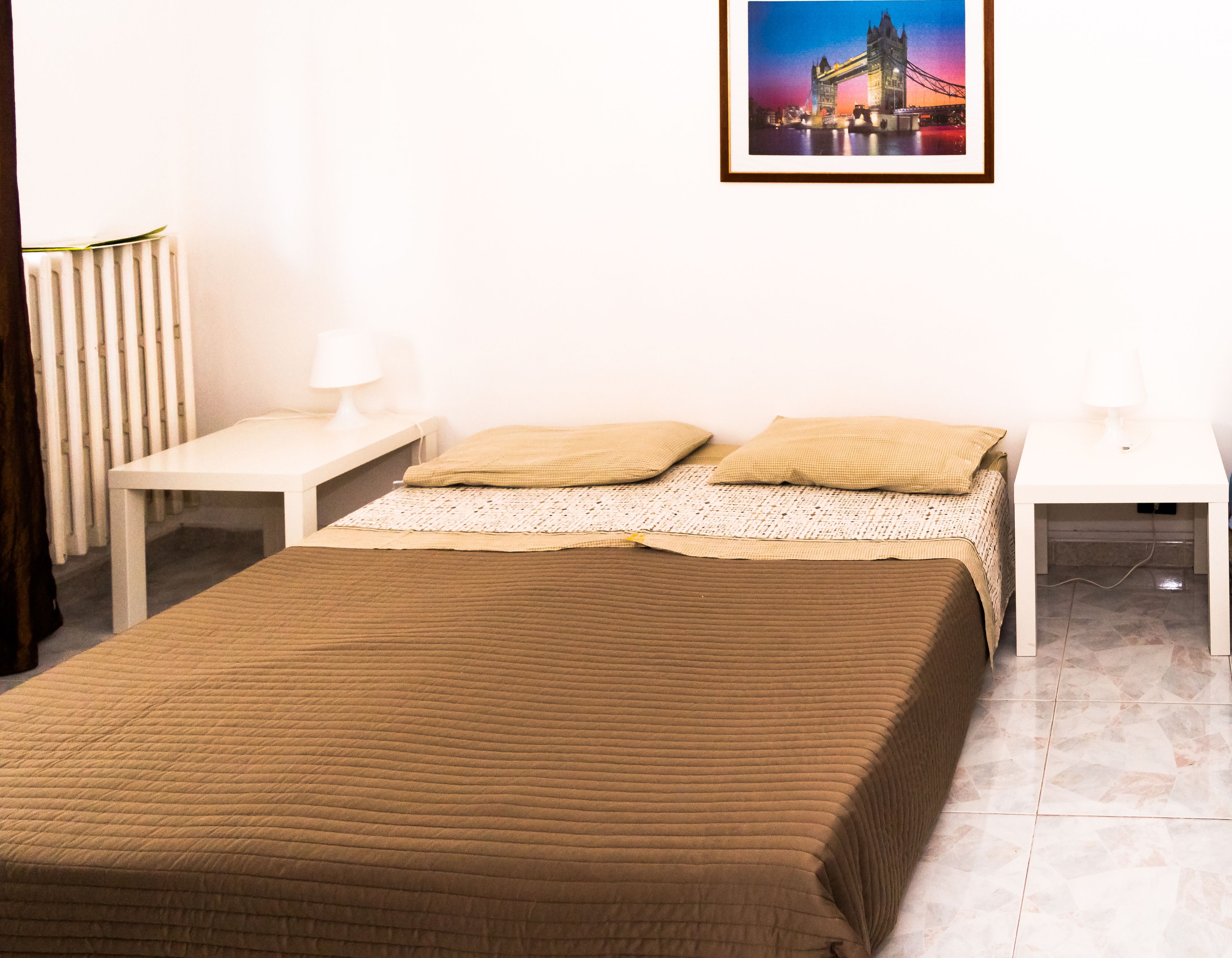 A neatly made bed with a brown quilt, two bedside tables with lamps, and a framed picture of a bridge on the wall.