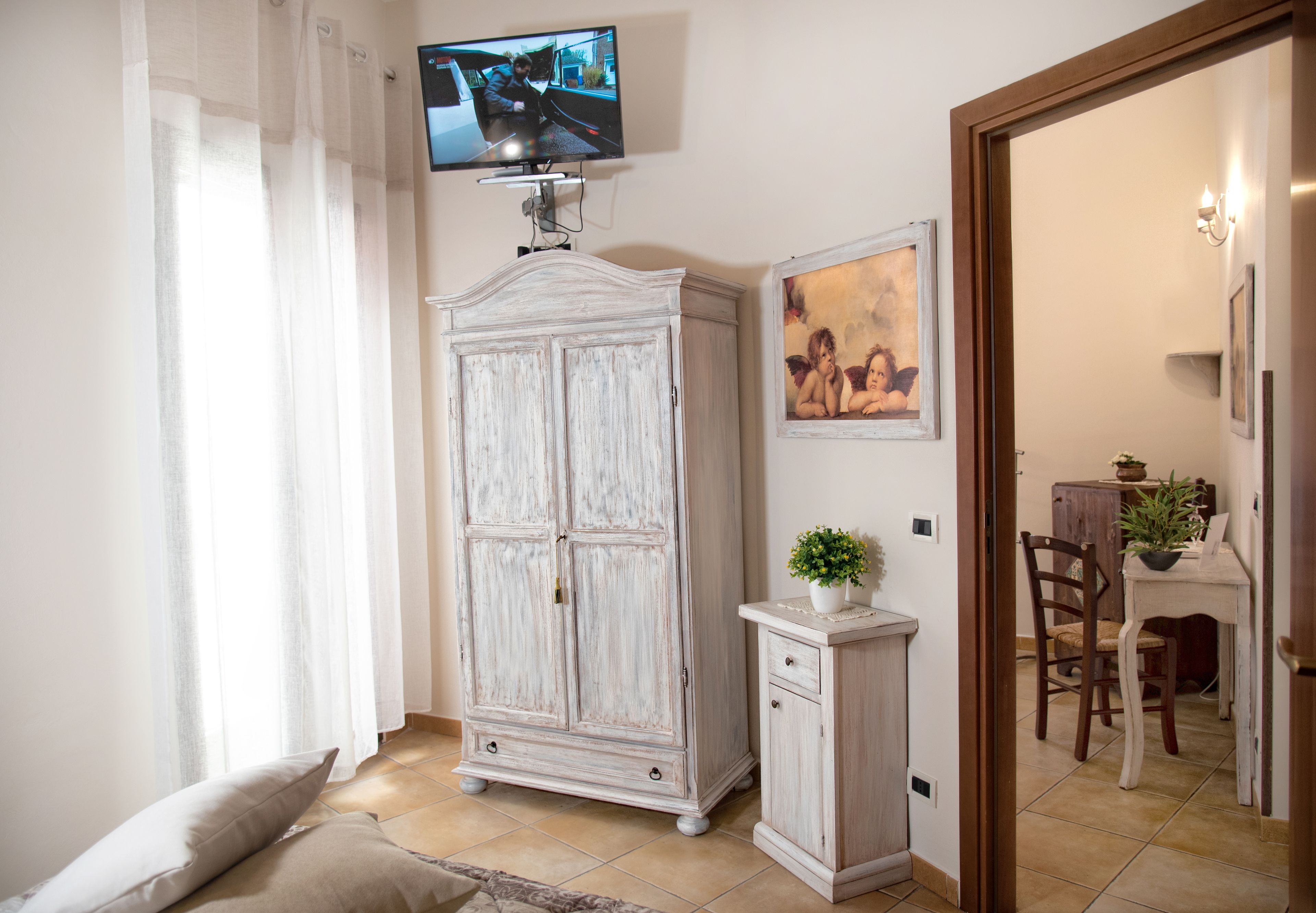 A room with a television mounted on the wall, a white wardrobe, a small cabinet, a framed picture, and a dining area visible through an open door.