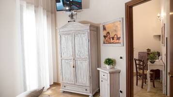 Una habitación con televisión montada en la pared, un armario blanco, un mueble pequeño, un cuadro enmarcado y una zona de comedor visible a través de una puerta abierta.