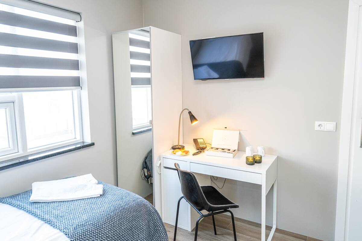 A bedroom with a bed, a desk with a chair, a TV, and a window with blinds.