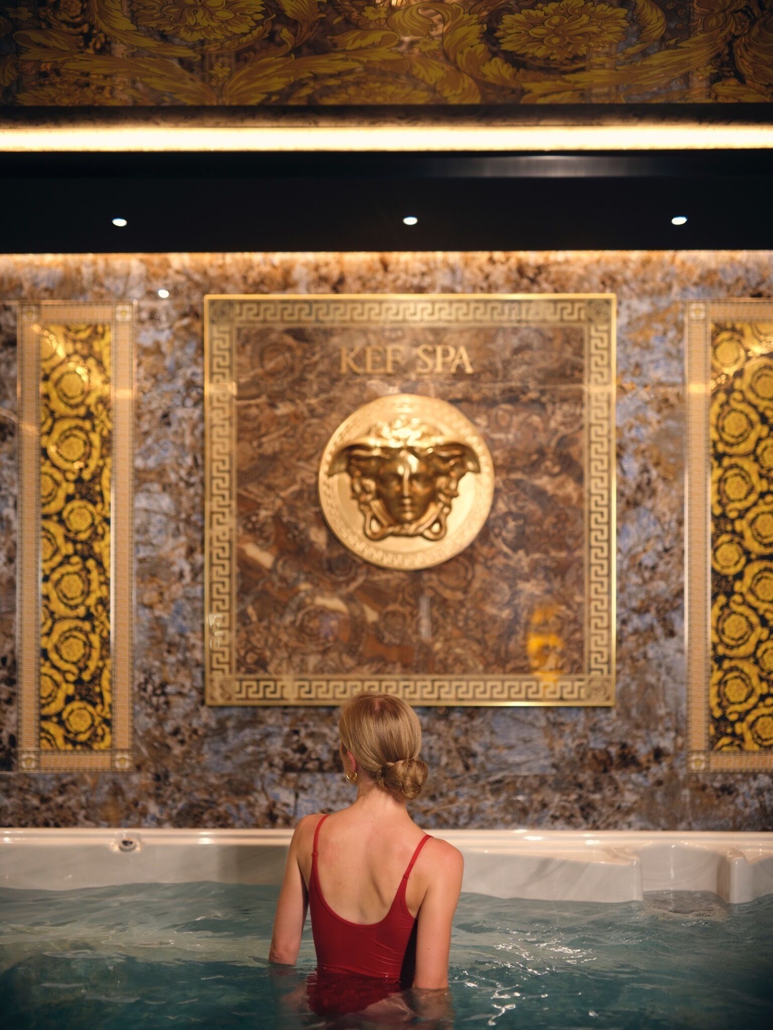 Eine Frau im roten Badeanzug in einem Spa-Pool mit einer Wandverkleidung, die ein zentrales Wappen und den Schriftzug "KEF SPA" zeigt.