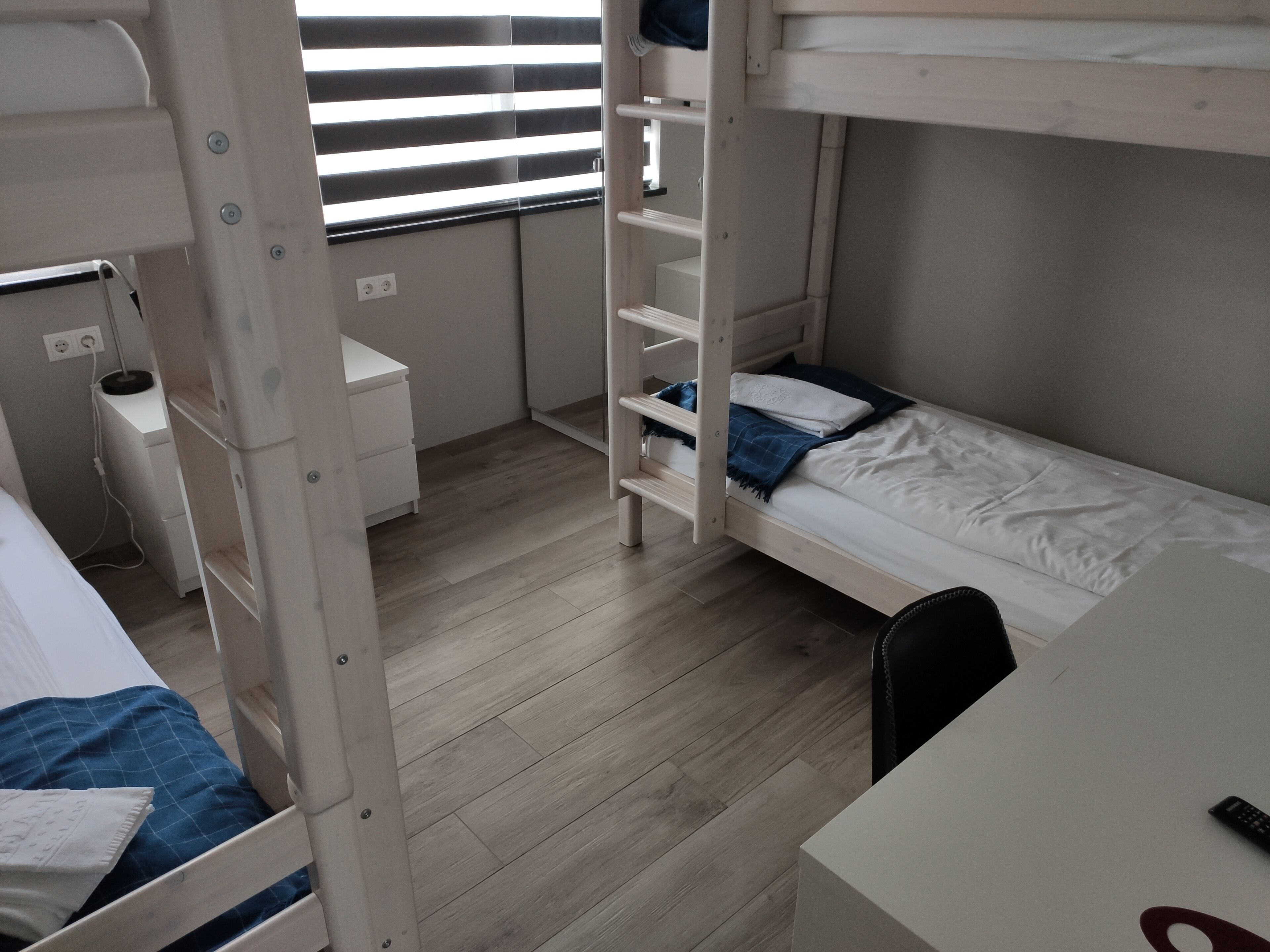 Une chambre avec un lit superposé, un bureau et une chaise, une petite table de chevet et une fenêtre avec des stores.