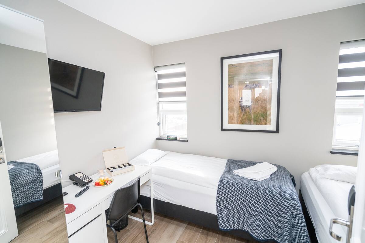 A hotel room with a bed, a desk with a chair, a flat-screen TV, and a window with blinds.