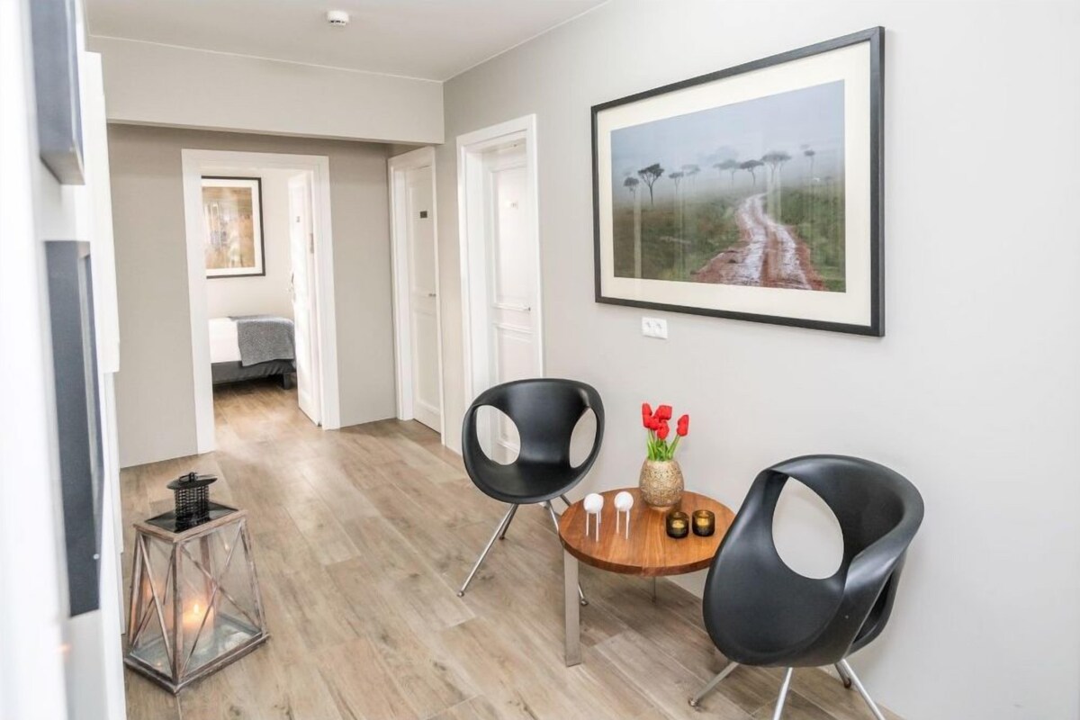 Uma sala de estar moderna com piso de madeira, duas cadeiras pretas, uma mesa redonda pequena com um vaso de flores vermelhas e um quadro de uma estrada com árvores na parede.