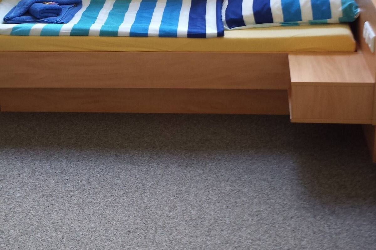 Una cama individual con un cojín a rayas y una toalla azul, un banco de madera y una ventana con cortinas.