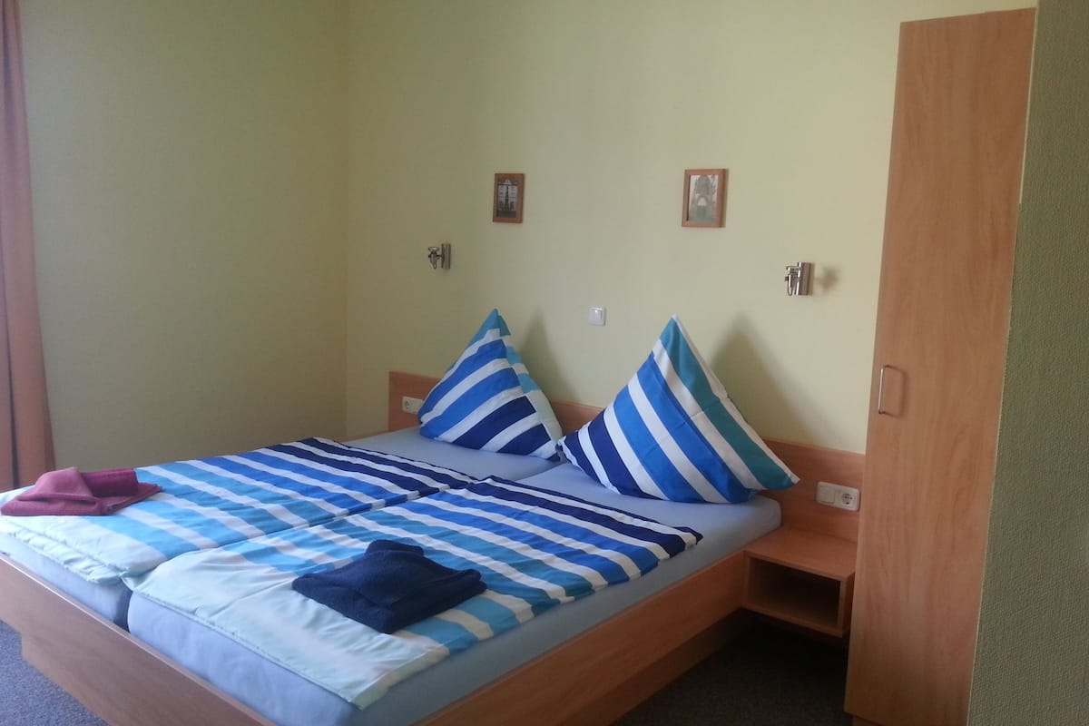 Una cama individual con una colcha a rayas y almohadas, un cabecero de madera, una mesita de noche pequeña y una puerta a la derecha.