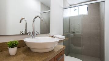 A modern bathroom with a large white sink, two faucets, a potted plant, a toilet, and a glass-enclosed shower.