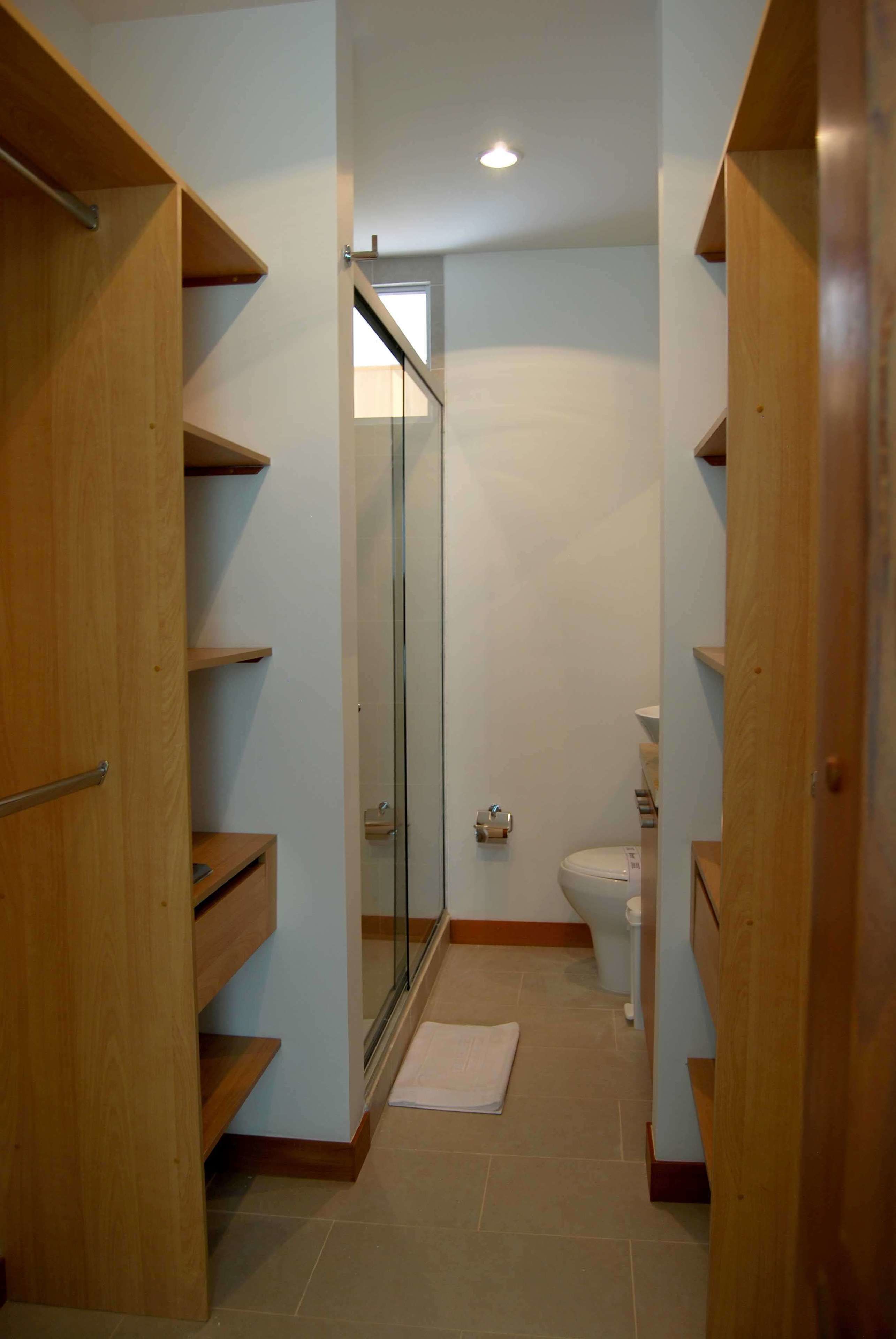 A narrow hallway with wooden shelving units on both sides, a glass door leading to a bathroom with a toilet and sink, and a small rug on the floor.