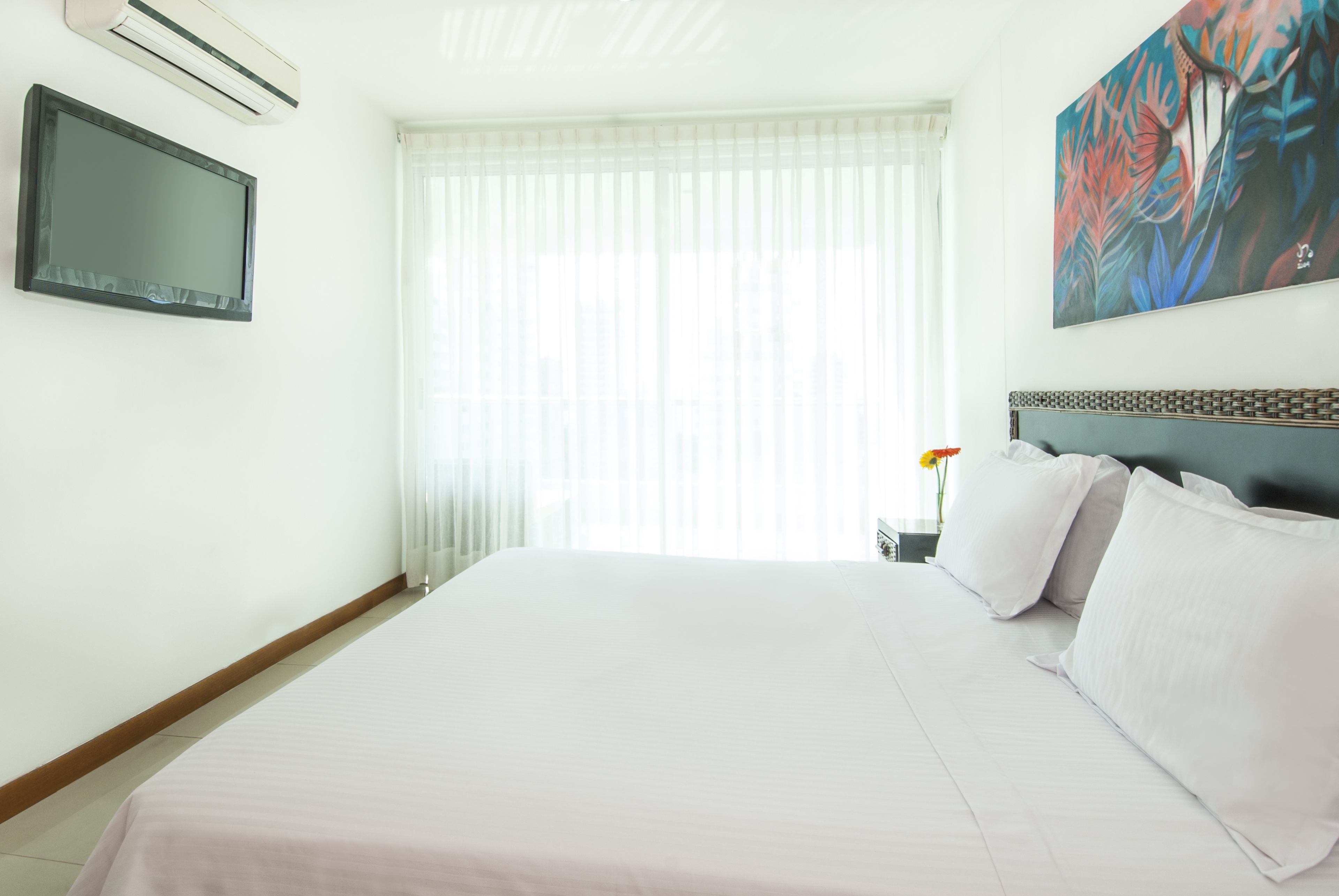 A hotel room with a large bed, a flat-screen TV, a painting, and a window with sheer curtains.
