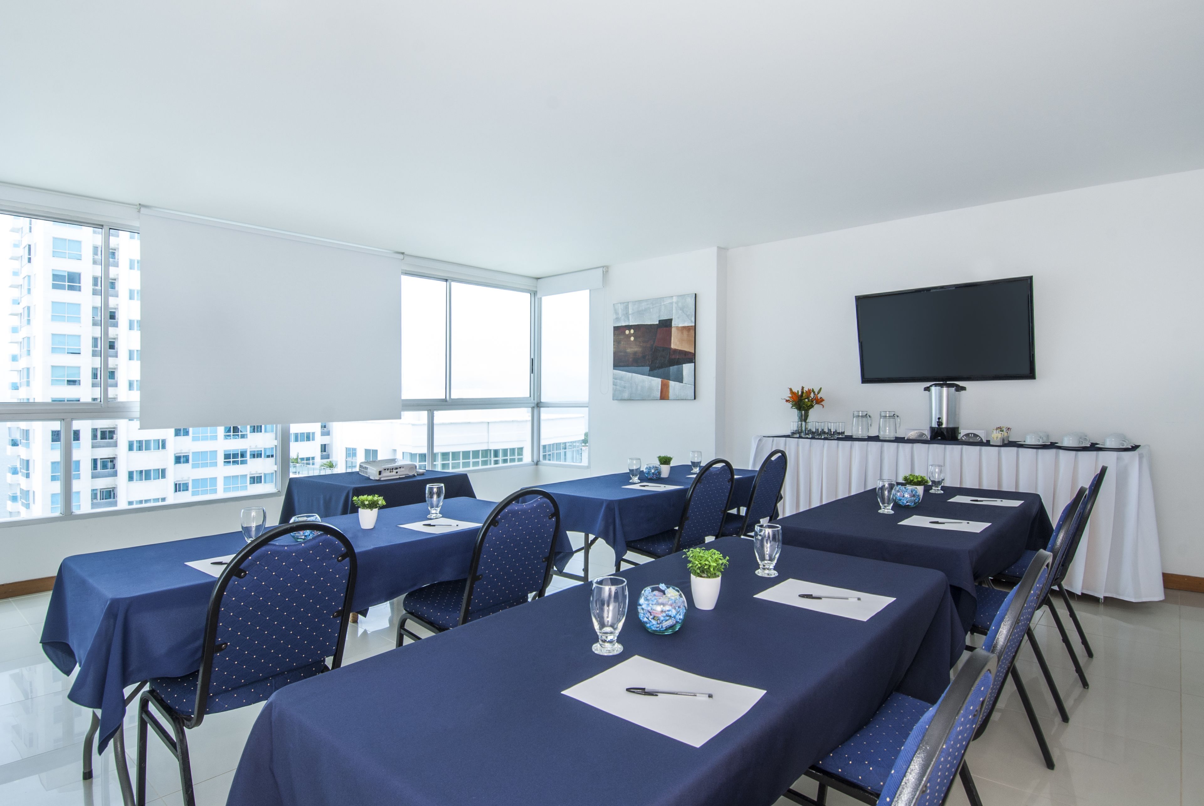 Une salle de conférence avec des tables disposées pour une réunion, une grande fenêtre donnant sur la ville et un téléviseur à écran plat fixé au mur.