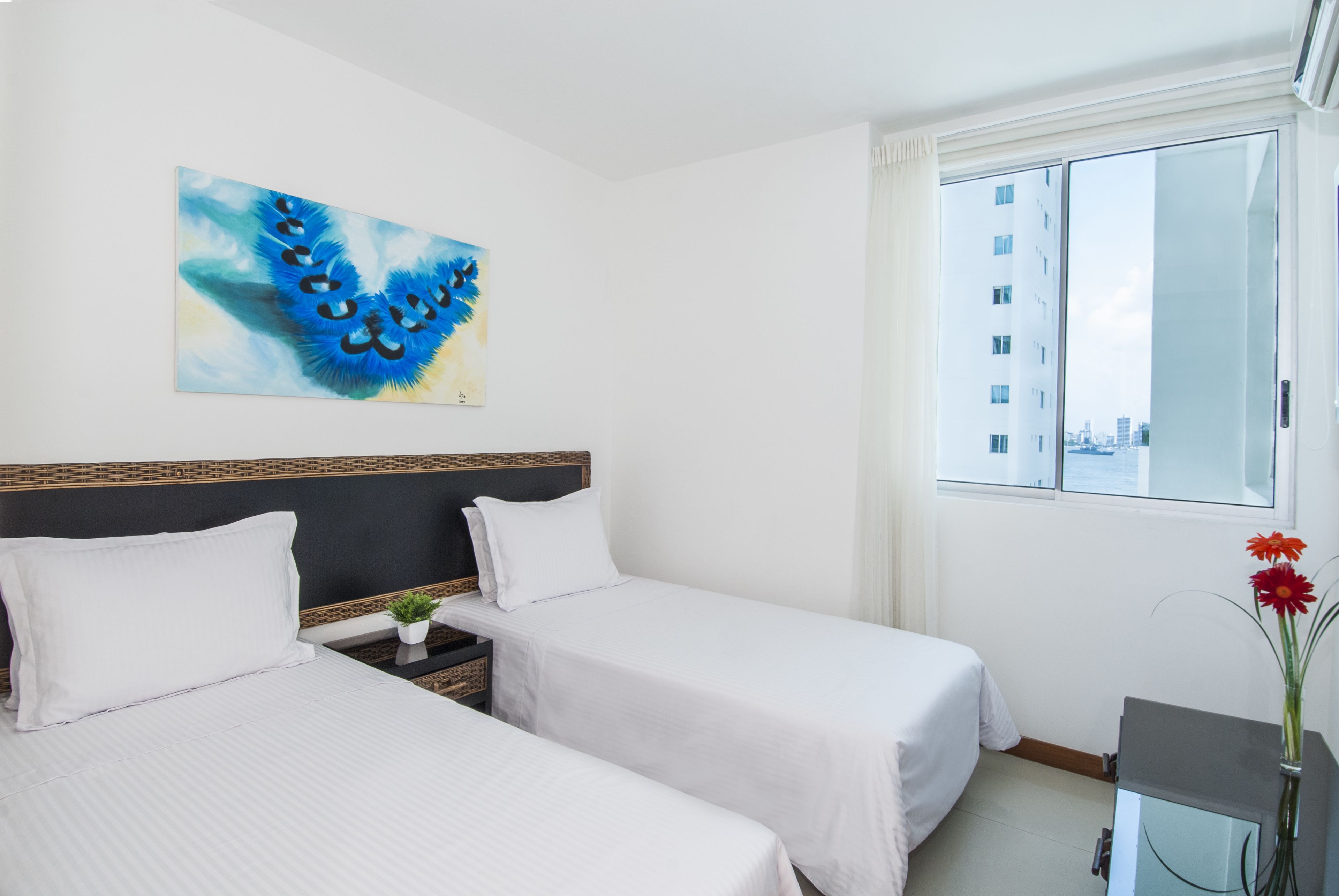 A hotel room with two beds, a large window with a city view, a painting on the wall, and a small table with a plant.