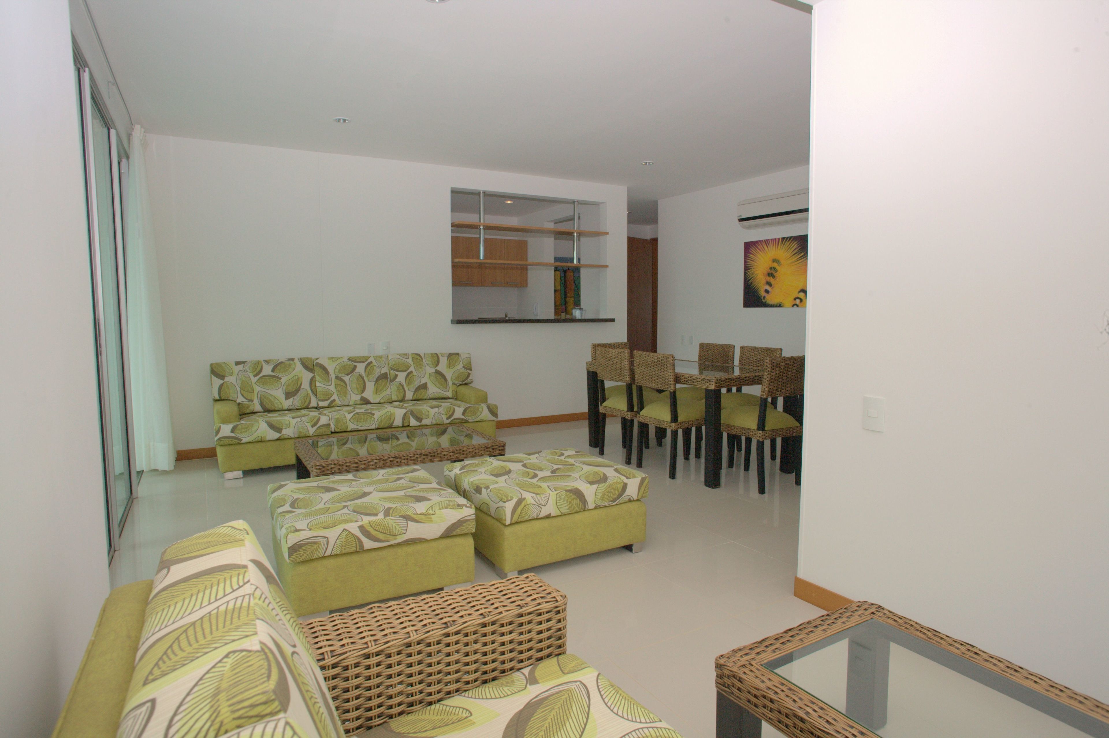 Una sala de estar con sofá, otomanas, una mesa de comedor con sillas y una cocina visible a través de un puerta de vidrio.