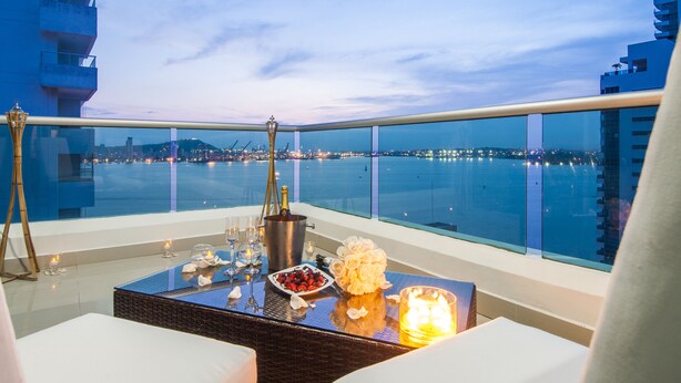 Un balcón con vistas al agua y las luces de la ciudad, una mesa con champán, flores y aperitivos, y una silla de mimbre.