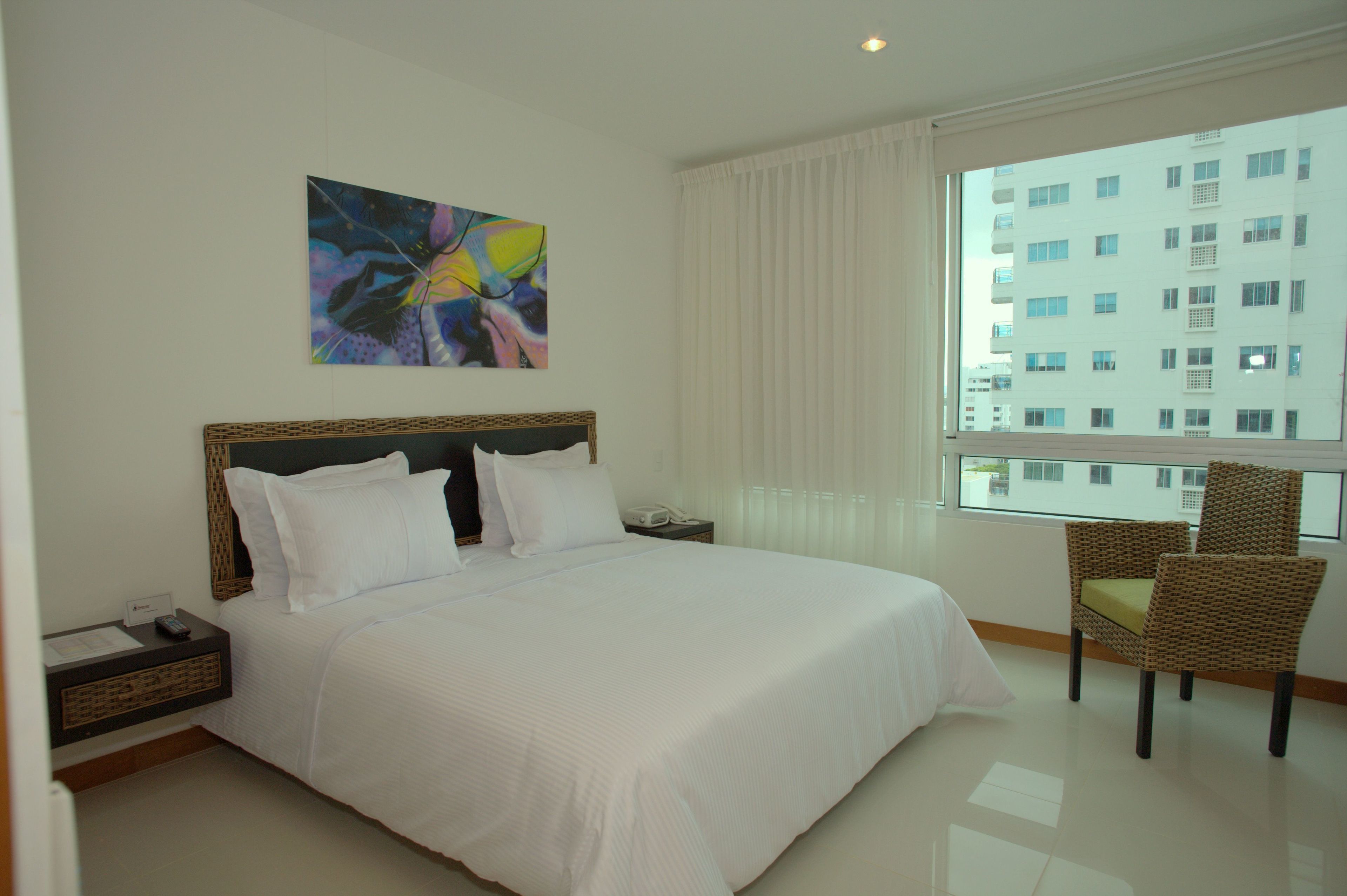A hotel room with a large bed, a nightstand, a chair, a painting on the wall, and a window with a view of buildings.