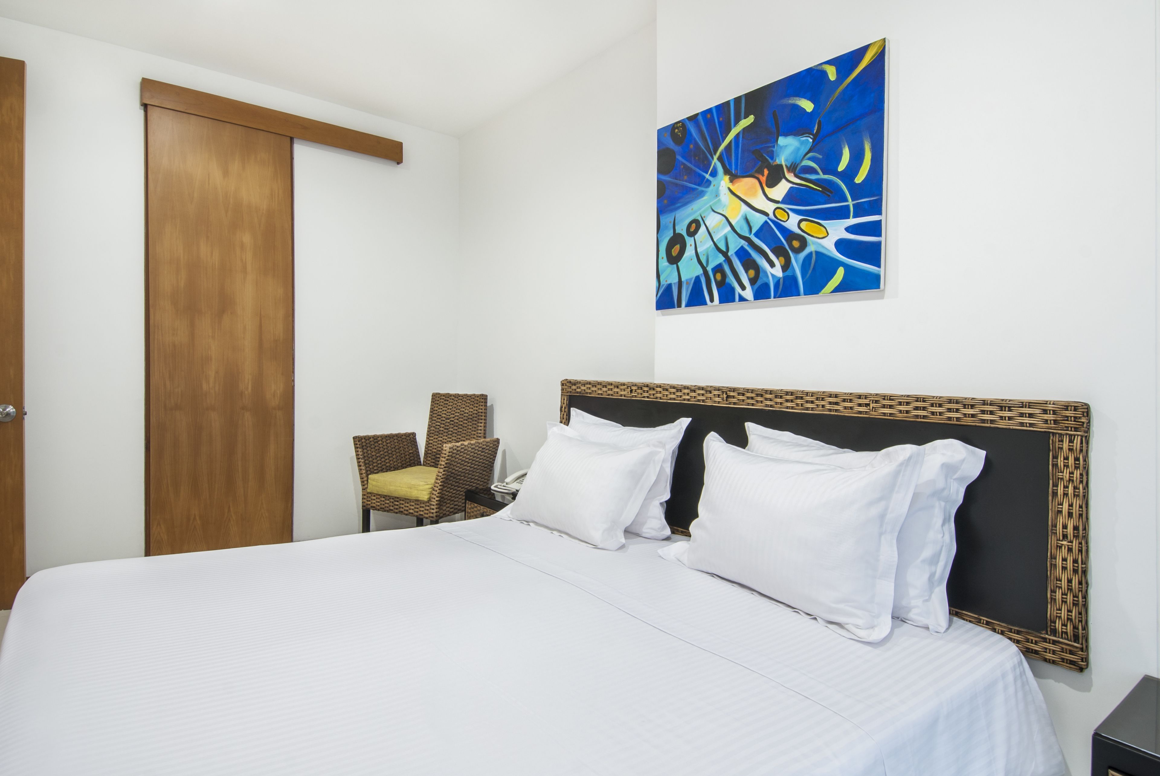 Habitación de hotel con una cama grande, un armario de madera, una silla de mimbre y un cuadro en la pared.