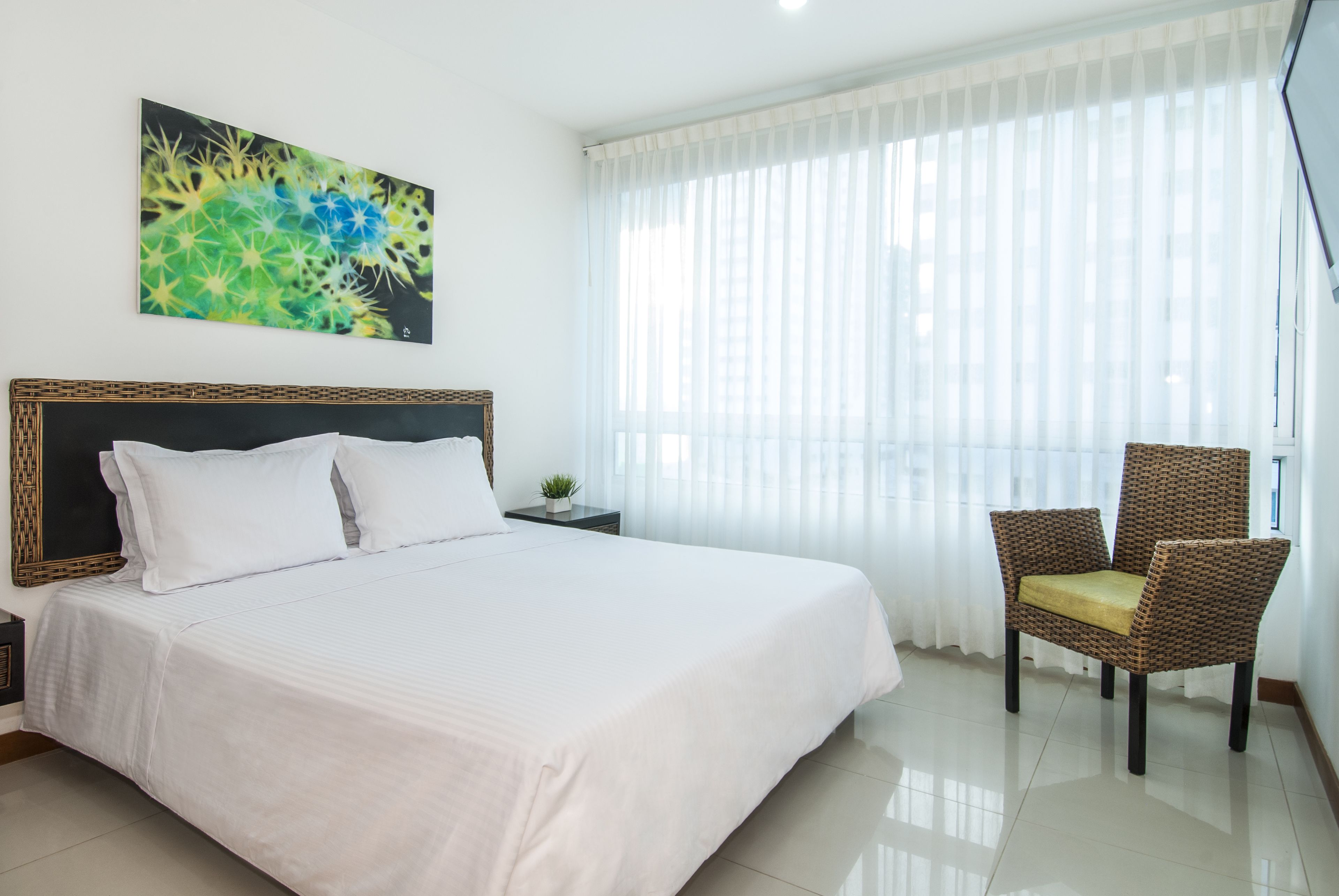 A hotel room with a large bed, a chair, a small table with a plant, and a painting on the wall.