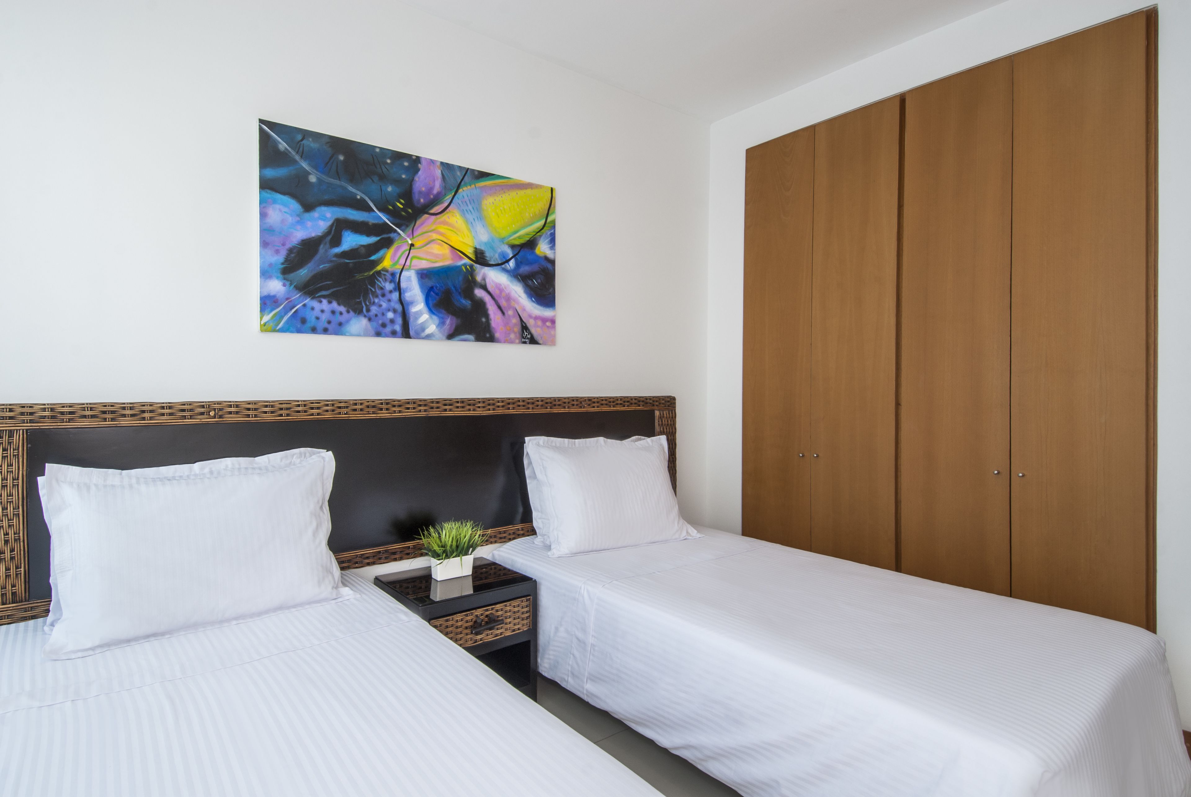 Habitación de hotel con dos camas, un armario de madera, una mesita de noche con una planta y un cuadro abstracto colorido en la pared.