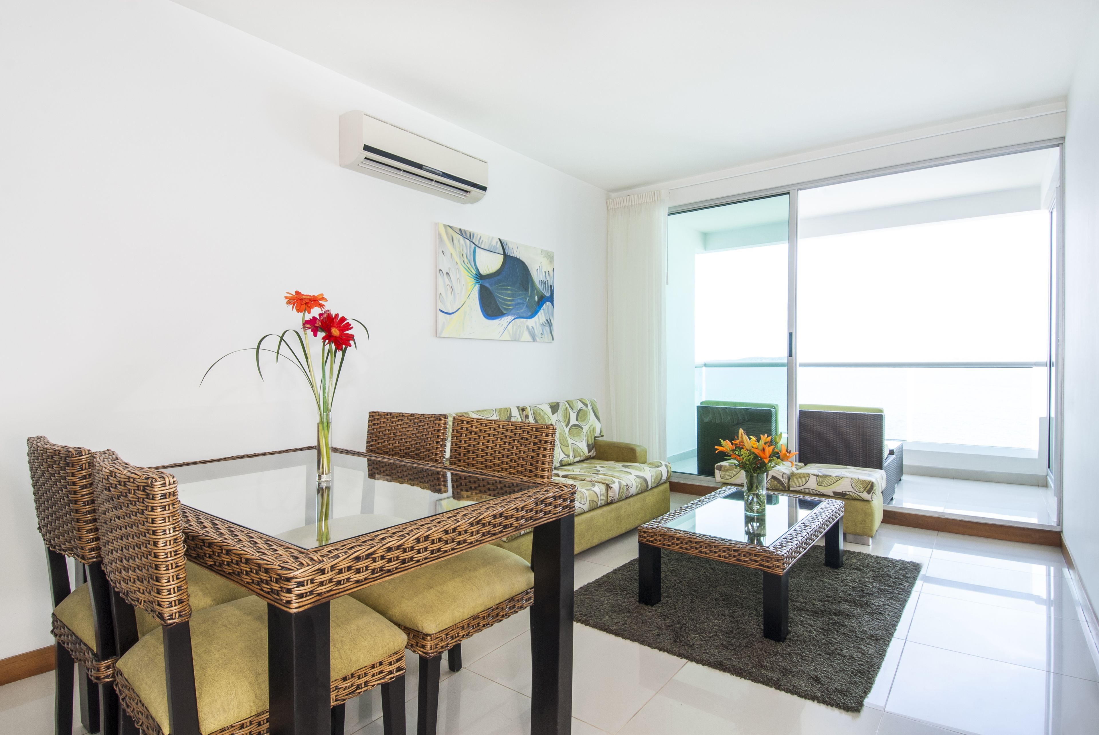 A modern living room with a glass coffee table, wicker chairs, a sofa, and a large window offering a view of the outdoors.