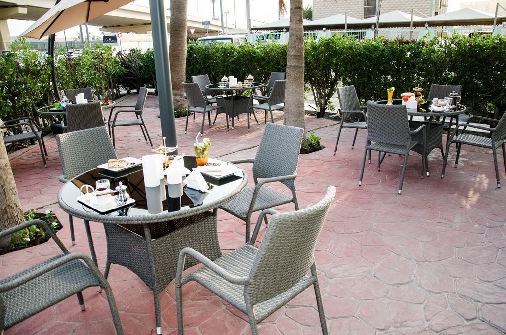 An outdoor patio with wicker tables and chairs, surrounded by greenery and a paved floor.