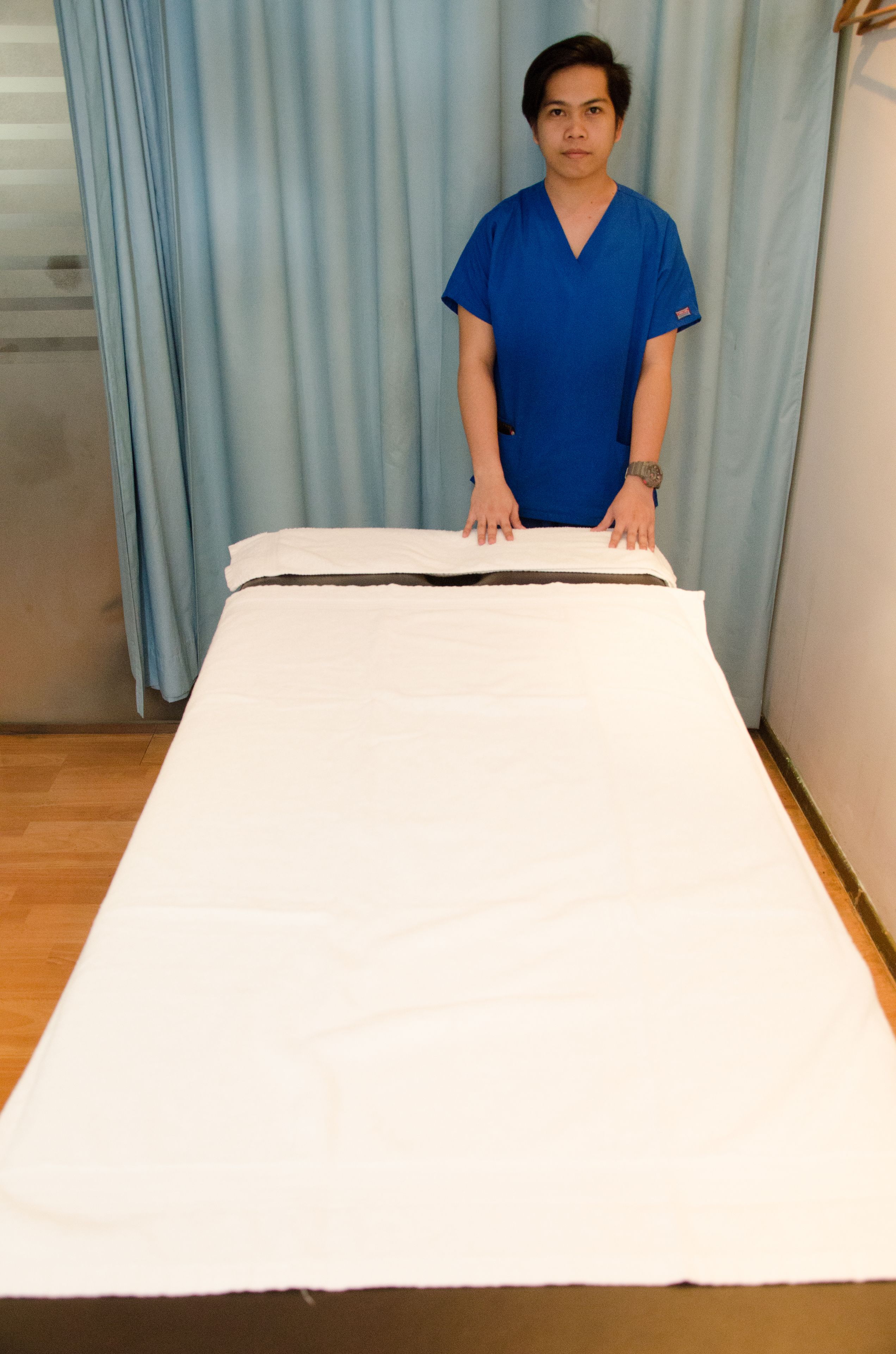 A person in blue scrubs standing behind a bed with a white sheet.