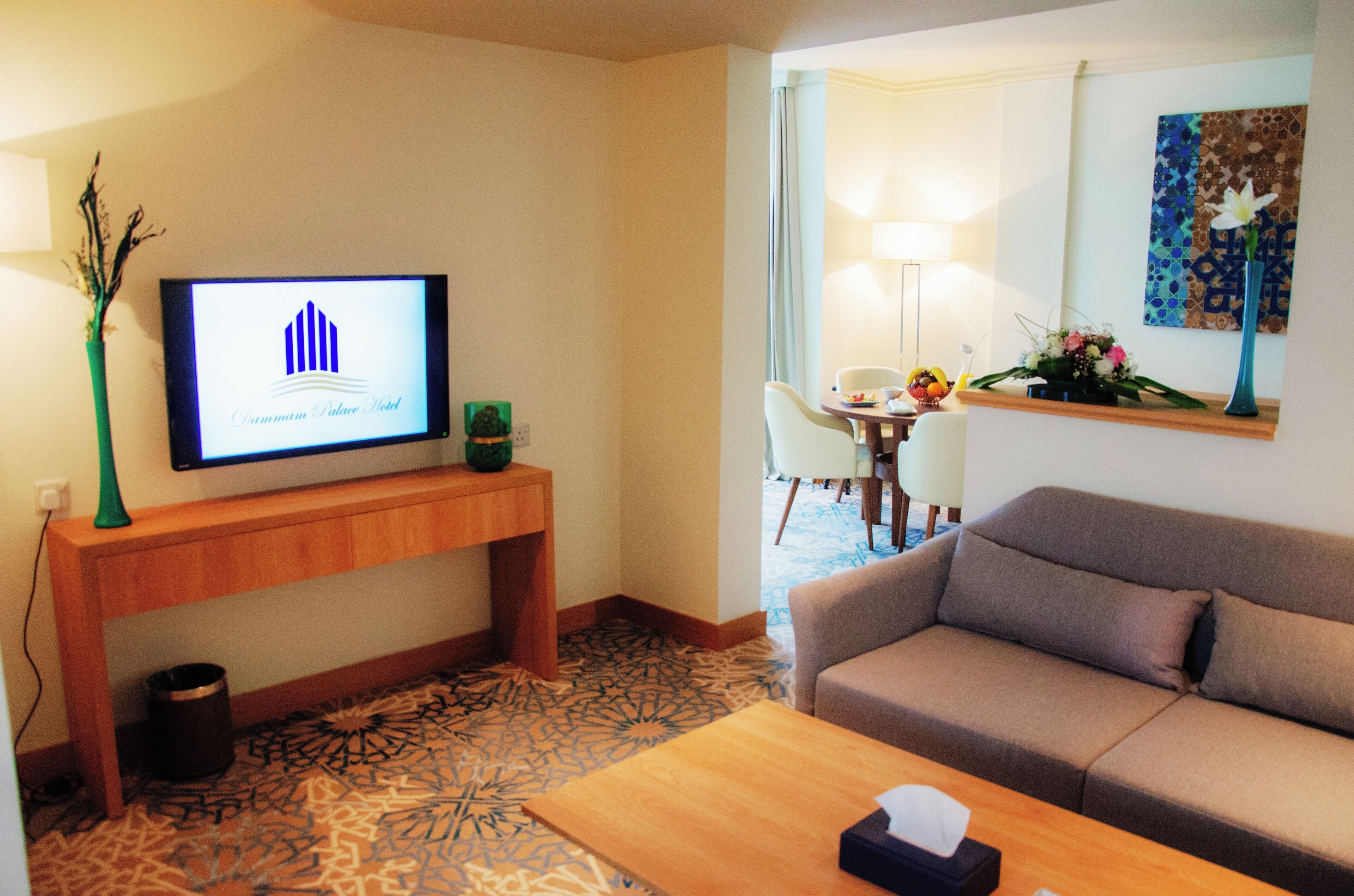 Habitación de hotel con televisor de pantalla plana sobre una consola de madera, un sofá, una mesa de centro y una alfombra estampada.