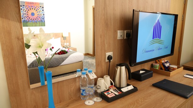 A hotel room with a wooden desk, a television displaying the hotel logo, a bed with a floral arrangement, and a mirror reflecting the room. - Dammam Palace Hotel (Dammam)