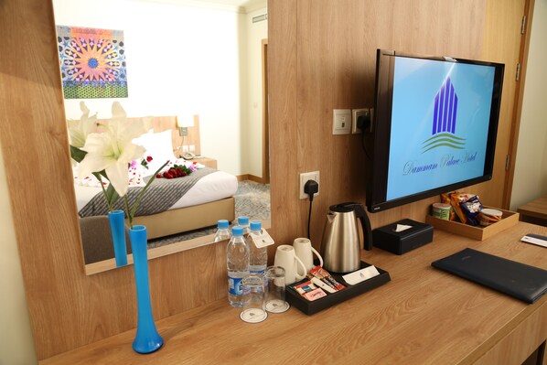 A hotel room with a wooden desk, a television displaying the hotel logo, a bed with a floral arrangement, and a mirror reflecting the room. - Dammam Palace Hotel (Dammam)