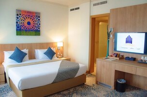 Quarto de hotel com uma cama, escrivaninha, computador e um objeto decorativo na parede.