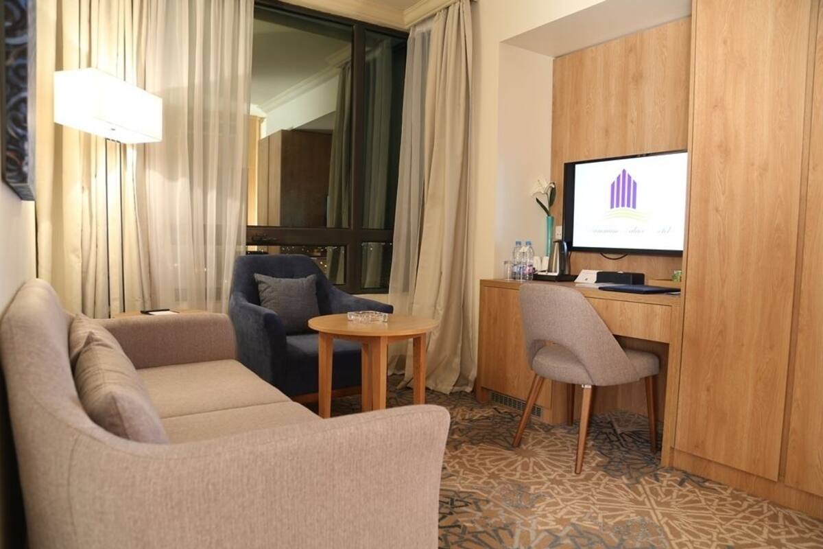 Quarto de hotel com sofá, uma cadeira, uma pequena mesa, escrivaninha com computador e televisão exibindo um gráfico.