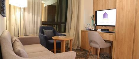 Quarto de hotel com sofá, uma cadeira, uma pequena mesa, escrivaninha com computador e televisão exibindo um gráfico.