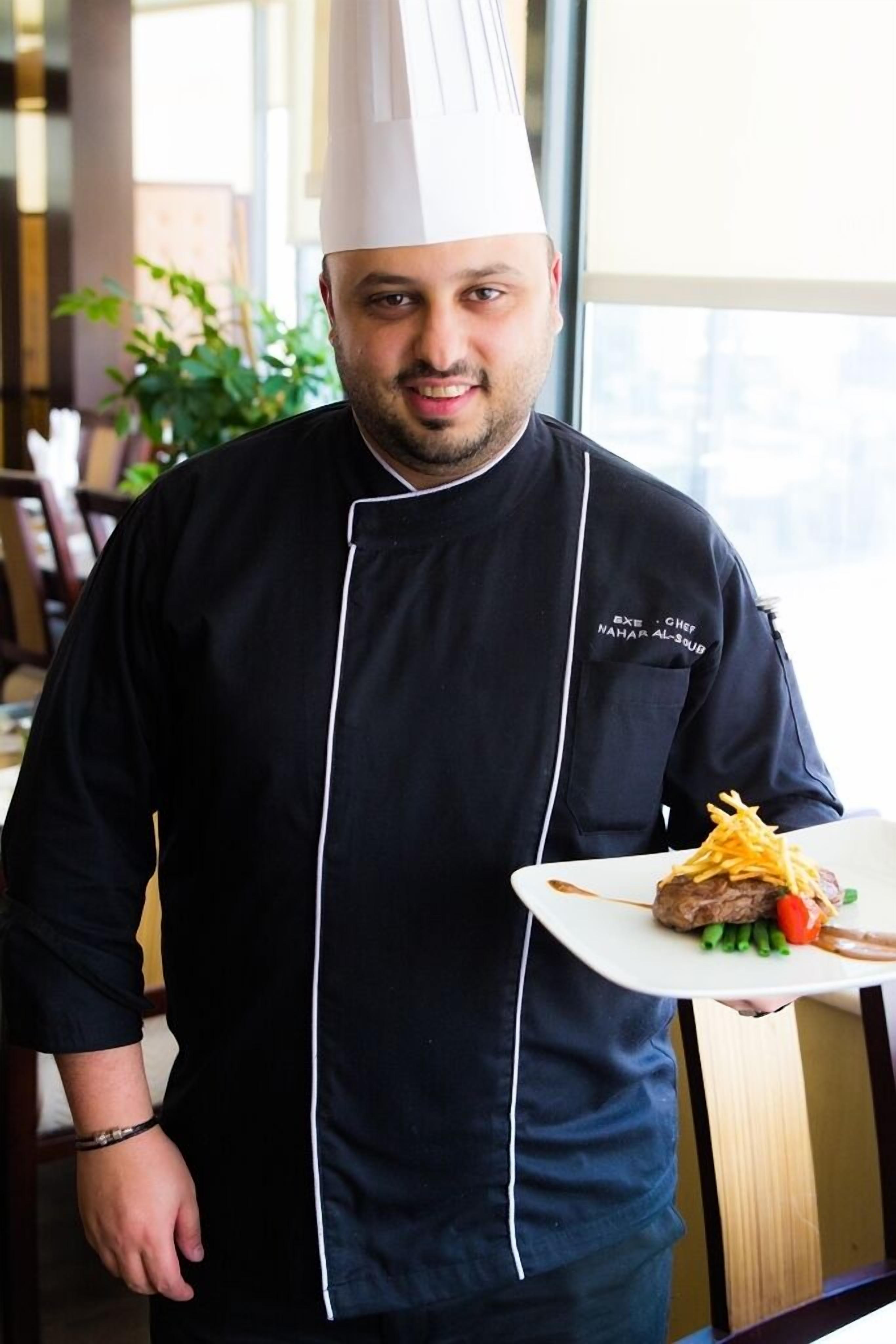 A chef in a black uniform with a white chef's hat, holding a plate with a grilled meat dish, green beans, and a slice of tomato.