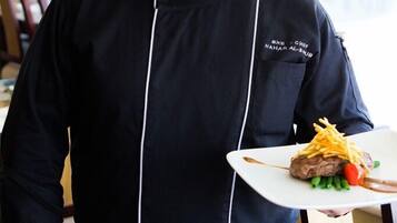 A chef in a black uniform with a white chef's hat, holding a plate with a grilled meat dish, green beans, and a slice of tomato.