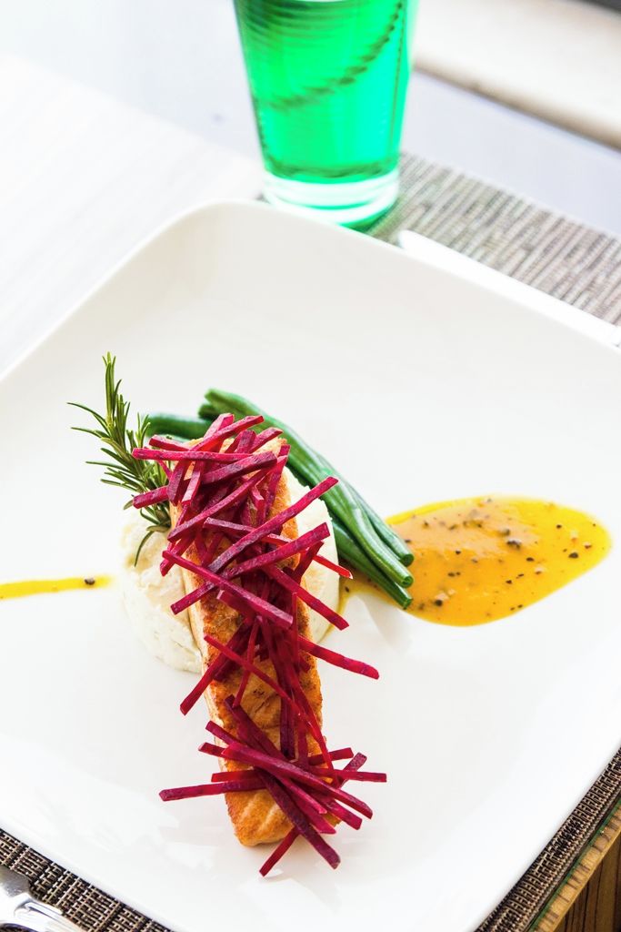 Un piatto quadrato con base bianca, un pesce guarnito con strisce di verdure rosse, fagiolini verdi e salsa gialla.