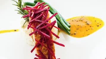 Prato quadrado com base branca, um pedaço de peixe coberto com tiras de vegetais vermelhos, feijão verde e um molho amarelo.