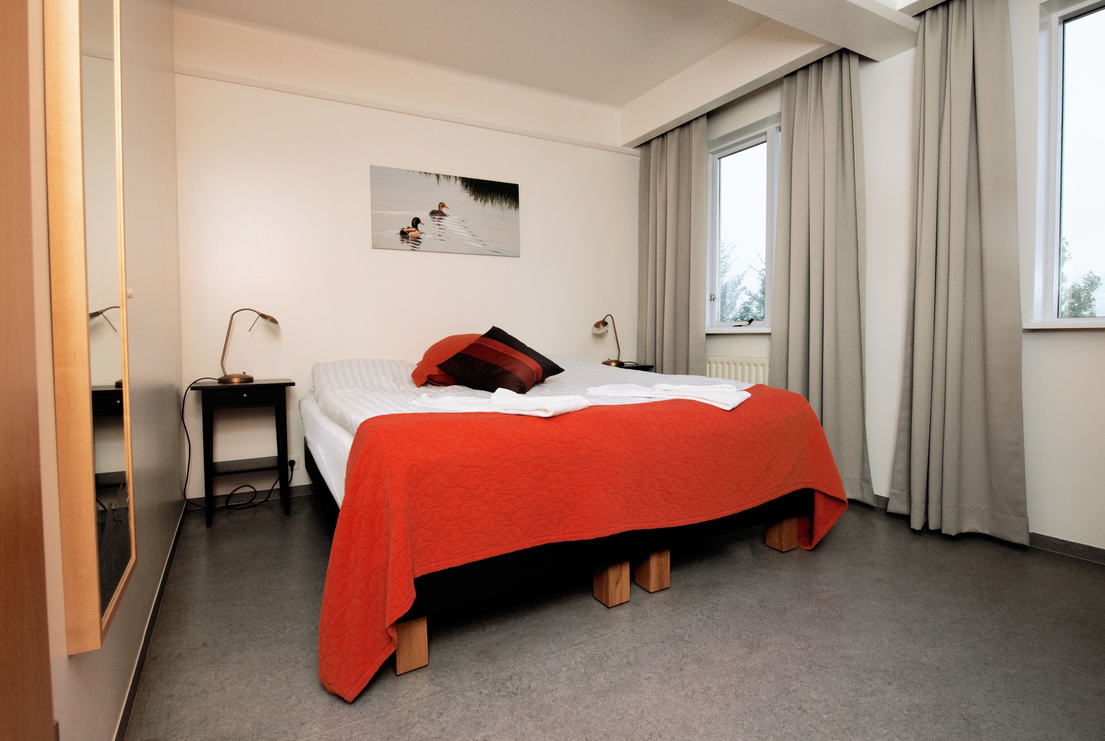 Ein Hotelzimmer mit Bett, Nachttisch, Lampe, einem Fenster mit Vorhängen und einem Gemälde an der Wand.