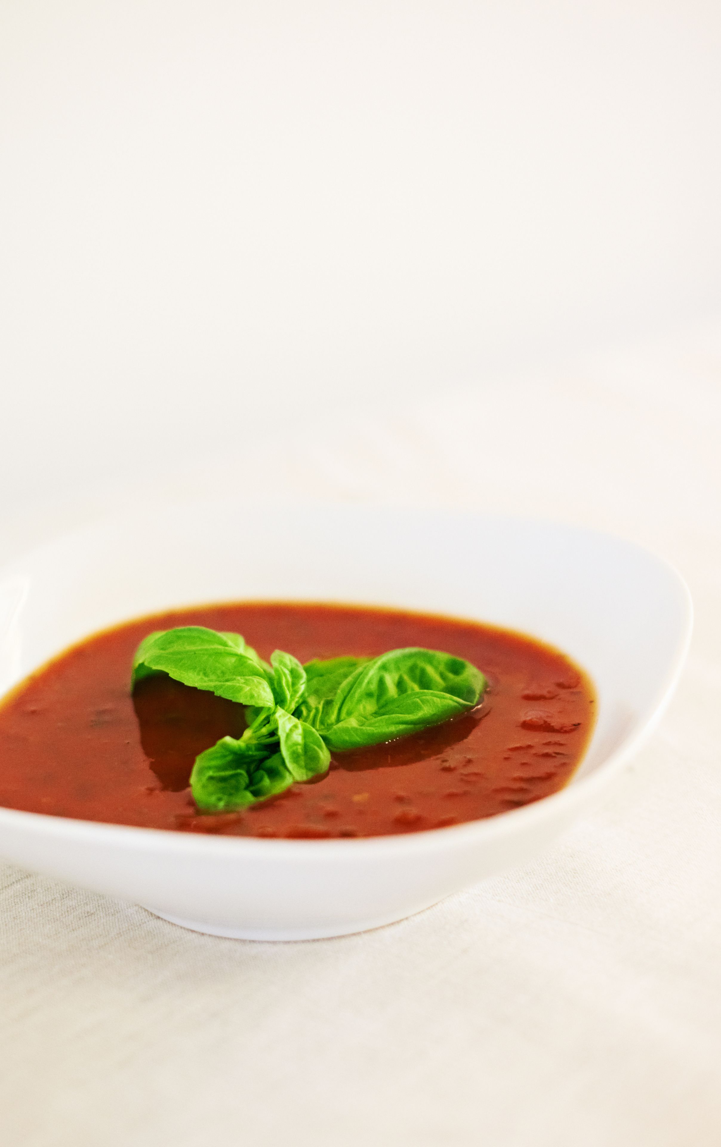 Un tazón blanco de sopa de tomate con hojas de albahaca sobre una superficie blanca.
