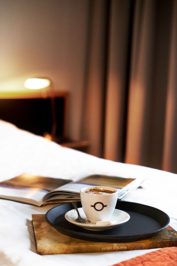 A cup of coffee on a tray with a spoon, an open book, and a bed with a white cover. - Hótel Varmahlíð (Skagafjörour)