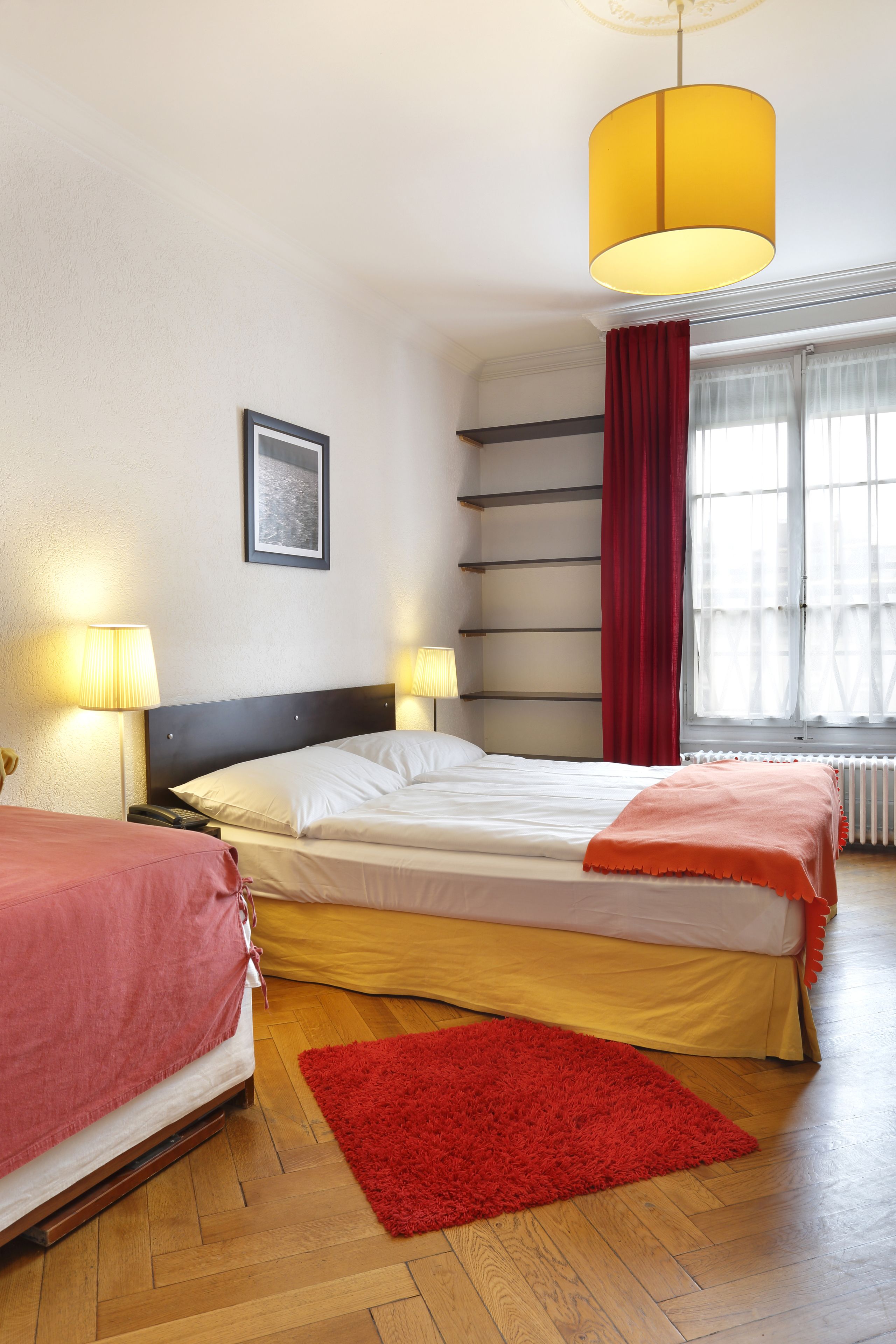 A hotel room with a double bed, a red rug, two bedside lamps, a window with curtains, and a picture on the wall.