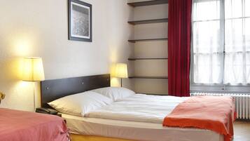 A hotel room with a double bed, a red rug, two bedside lamps, a window with curtains, and a picture on the wall.