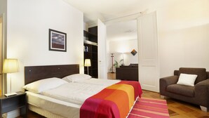 A hotel room with a bed, a nightstand, a lamp, a chair, a bookshelf, and a door leading to another room. - Torhôtel Genève (Geneva)