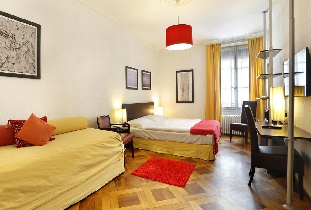 Habitación de hotel con dos camas, un escritorio, una silla, un tapete rojo y obras de arte en las paredes.