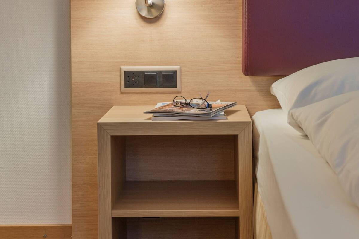 Ein ordentlich eingerichtetes Hotelzimmer mit einem Nachttisch aus Holz, einer Lampe, einem Bett mit weißer Bettwäsche und einem roten Kopfteil.