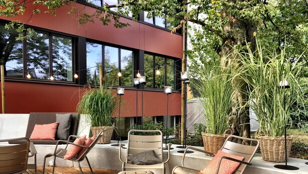 Een terras met moderne stoelen en een bank, omgeven door grote ramen en groen.
